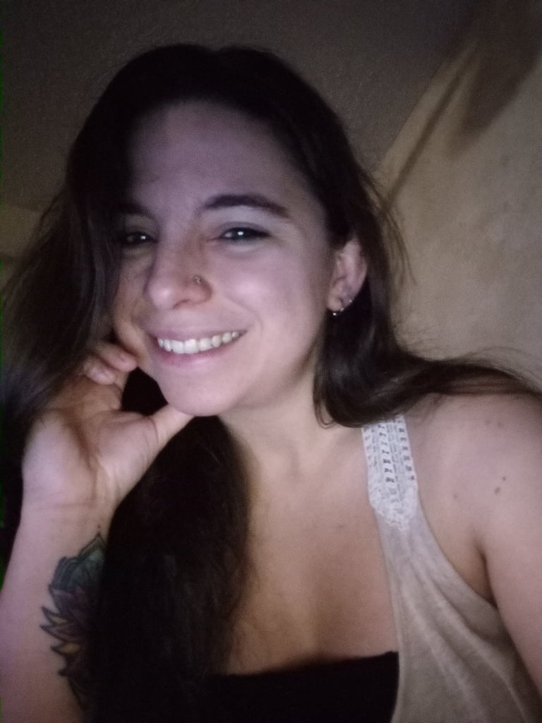Young woman smiling at camera with hand near face, wearing white tank top, dark background